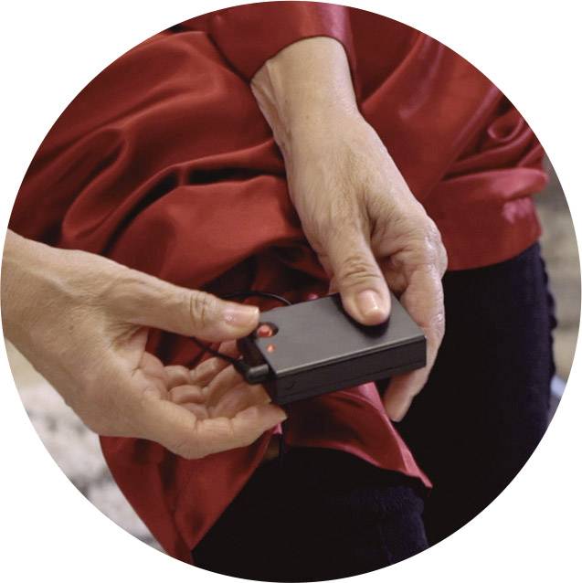 Une personne tient un petit appareil noir avec une lumière rouge, connecté à un fil, dans sa main droite, vêtue d'un tissu rouge.