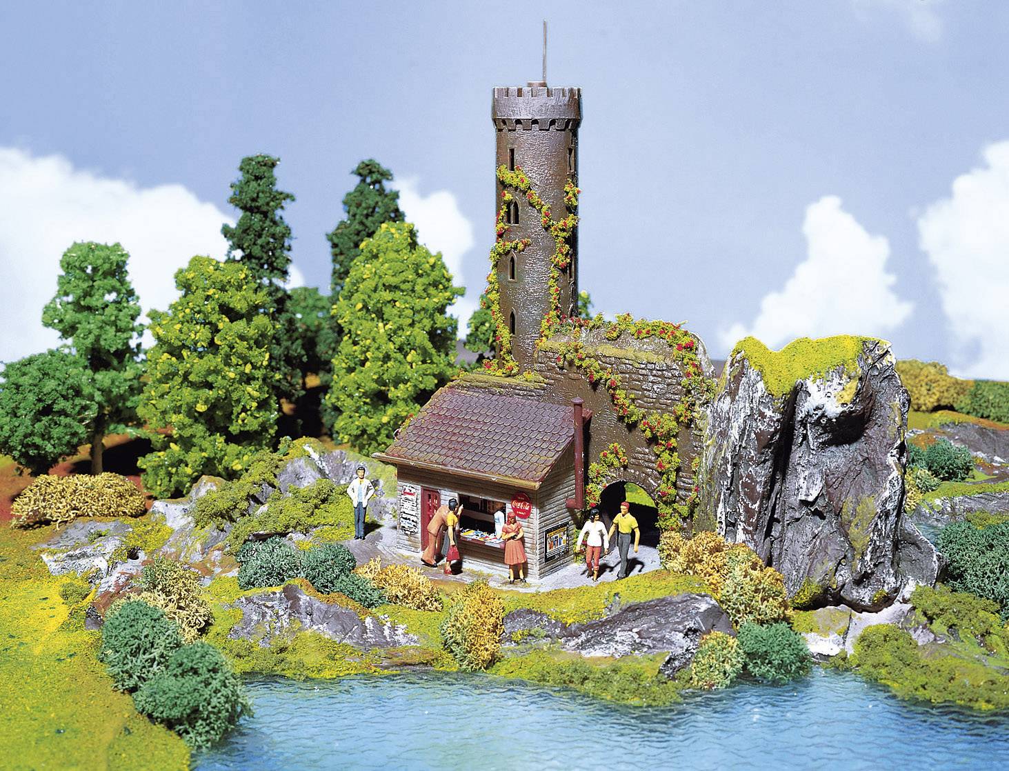 Un modèle miniature montre un château médiéval avec une tour, entouré d'arbres et d'un étang. Des figurines animent la scène.