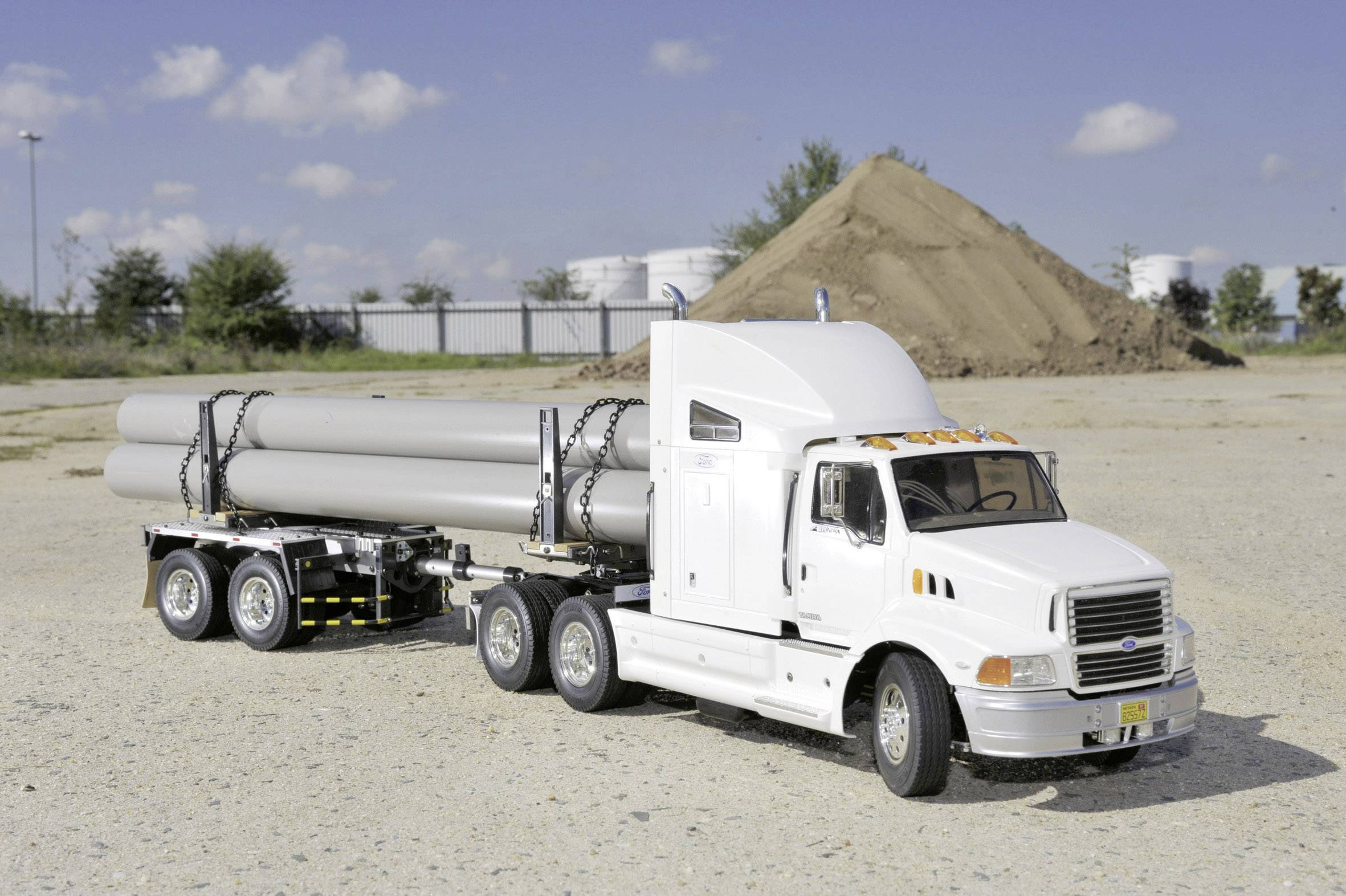 Un camion blanc avec une remorque, chargé de grands tuyaux, est stationné sur un terrain de gravier. À l'arrière-plan, on aperçoit un monticule de terre.