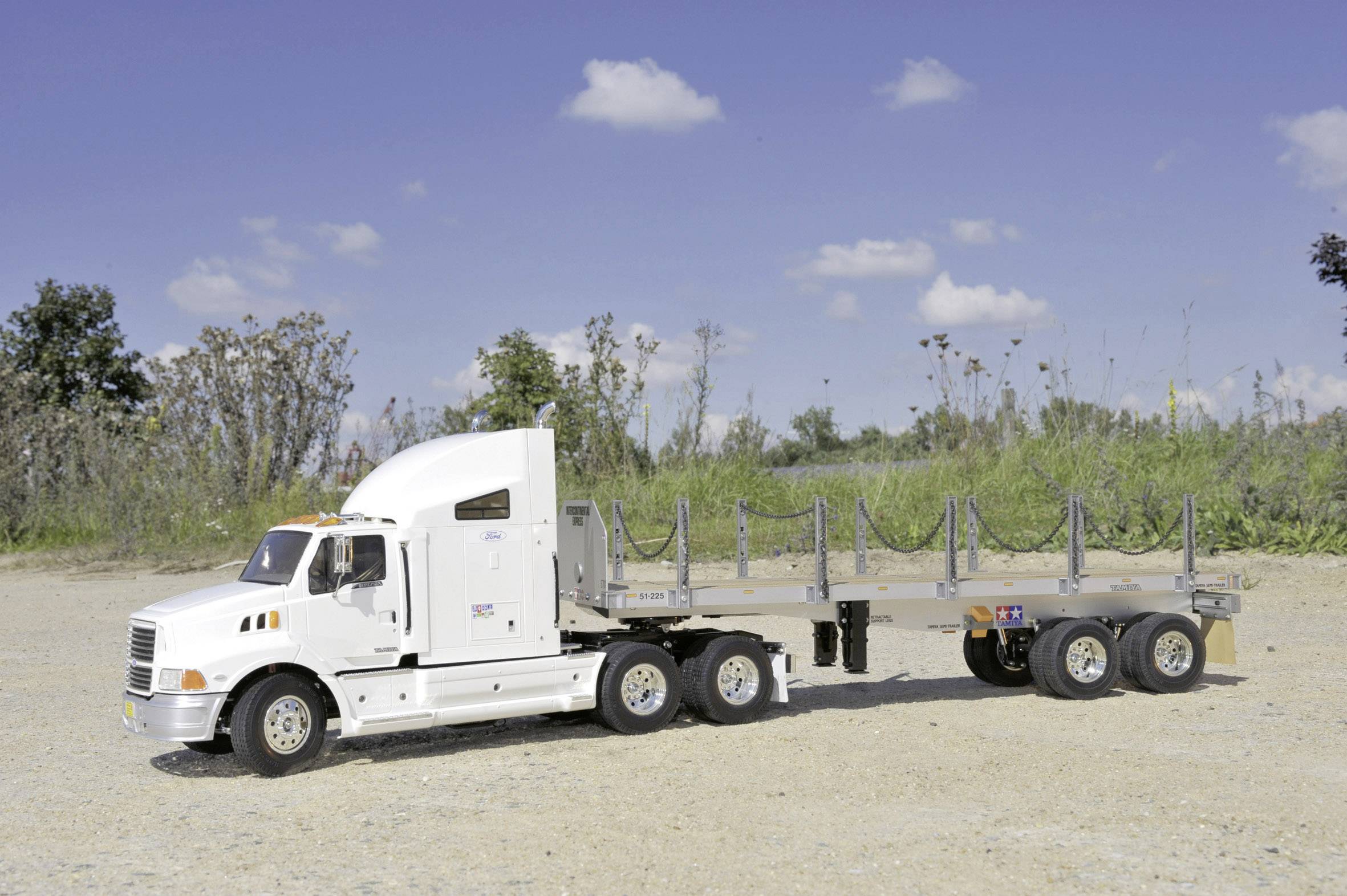 Un camion blanc avec une remorque est stationné sur un chemin non revêtu. Des arbres et un ciel bleu sont visibles en arrière-plan.