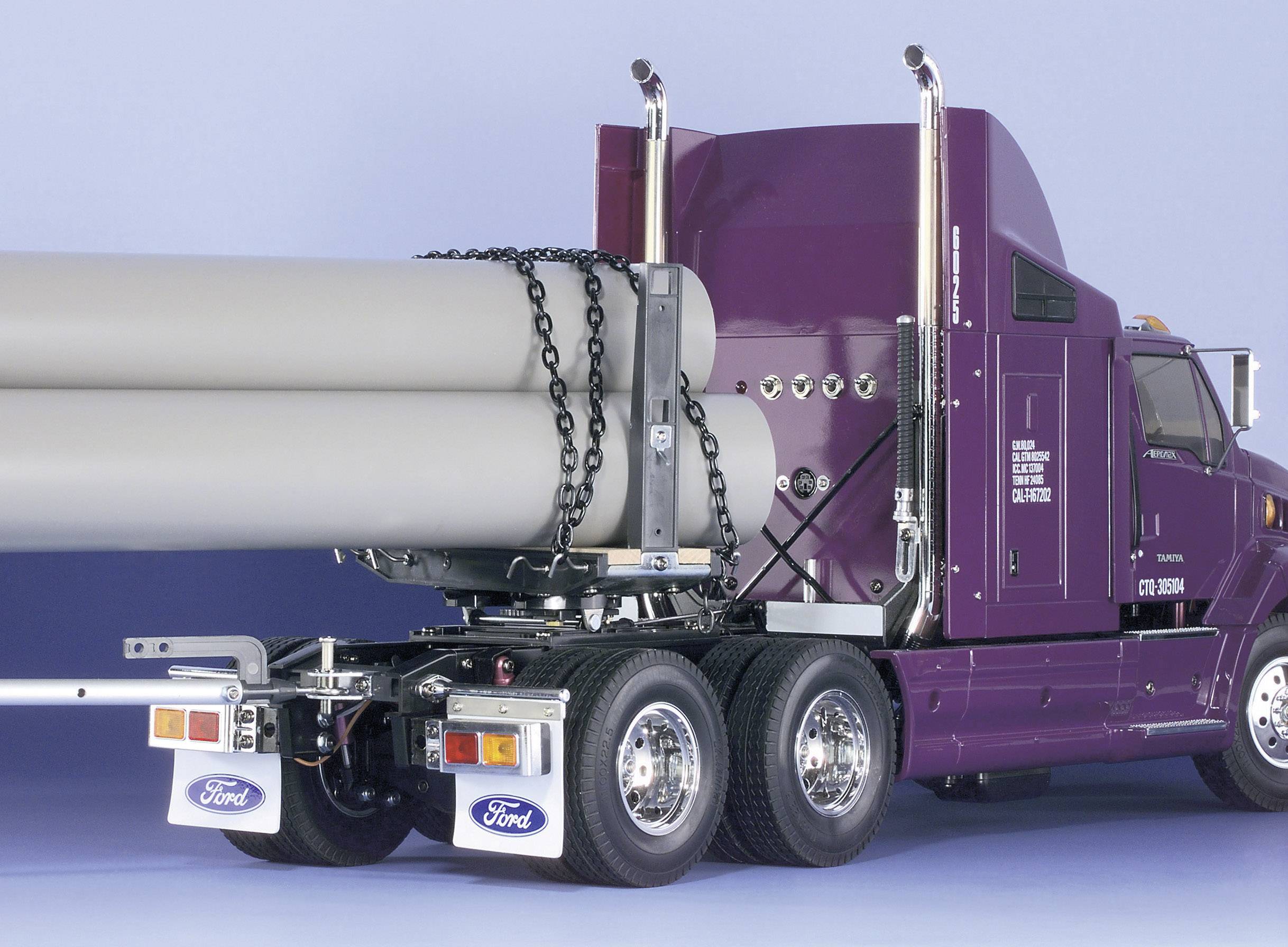 Un camion lilas transporte de grands tuyaux gris sécurisés par des chaînes. L'arrière-plan est d'un bleu clair uni.