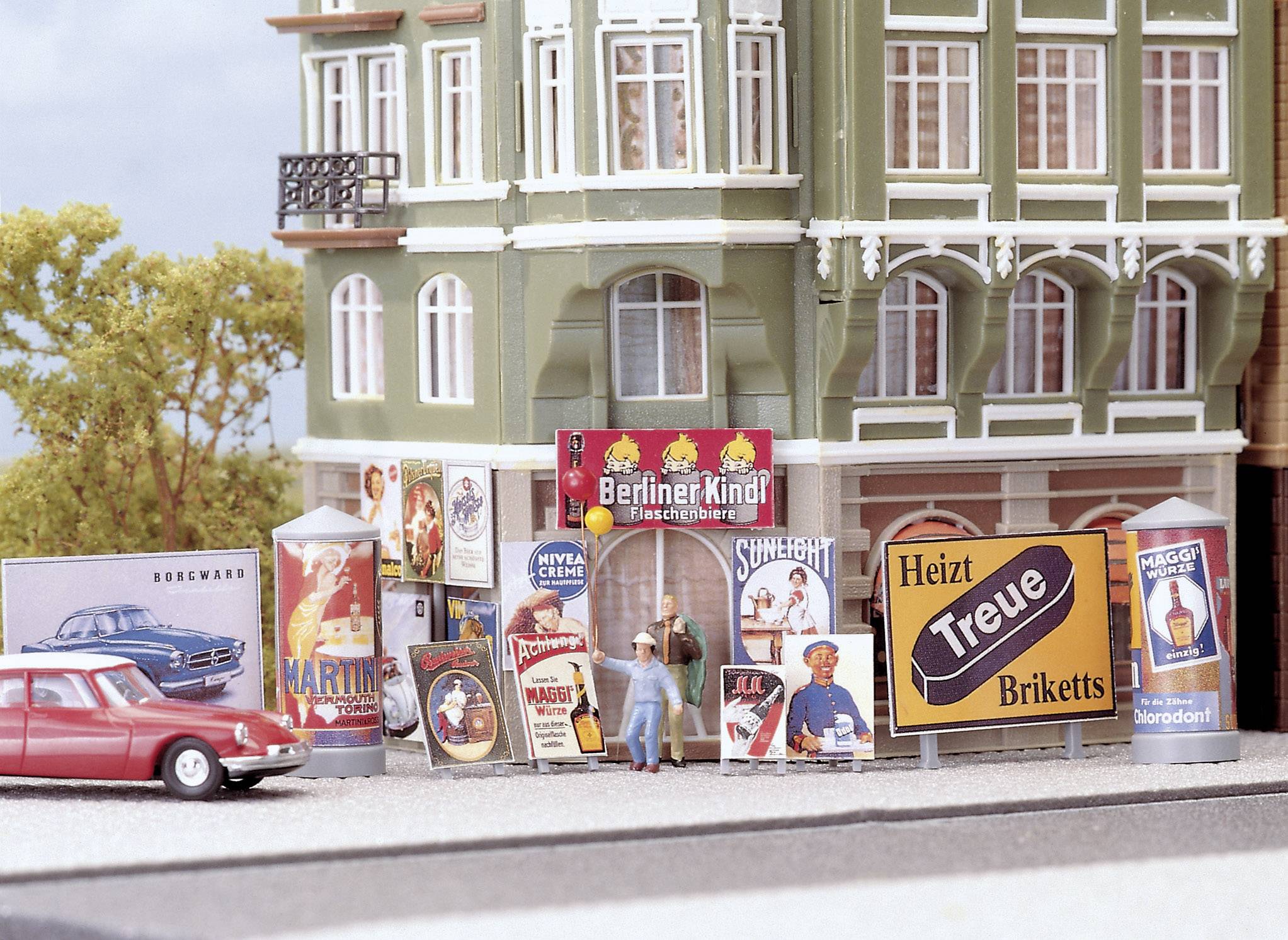 Un modèle d'un coin de rue avec des affiches publicitaires historiques sur un bâtiment. Une petite voiture de jouet rouge est garée à côté.