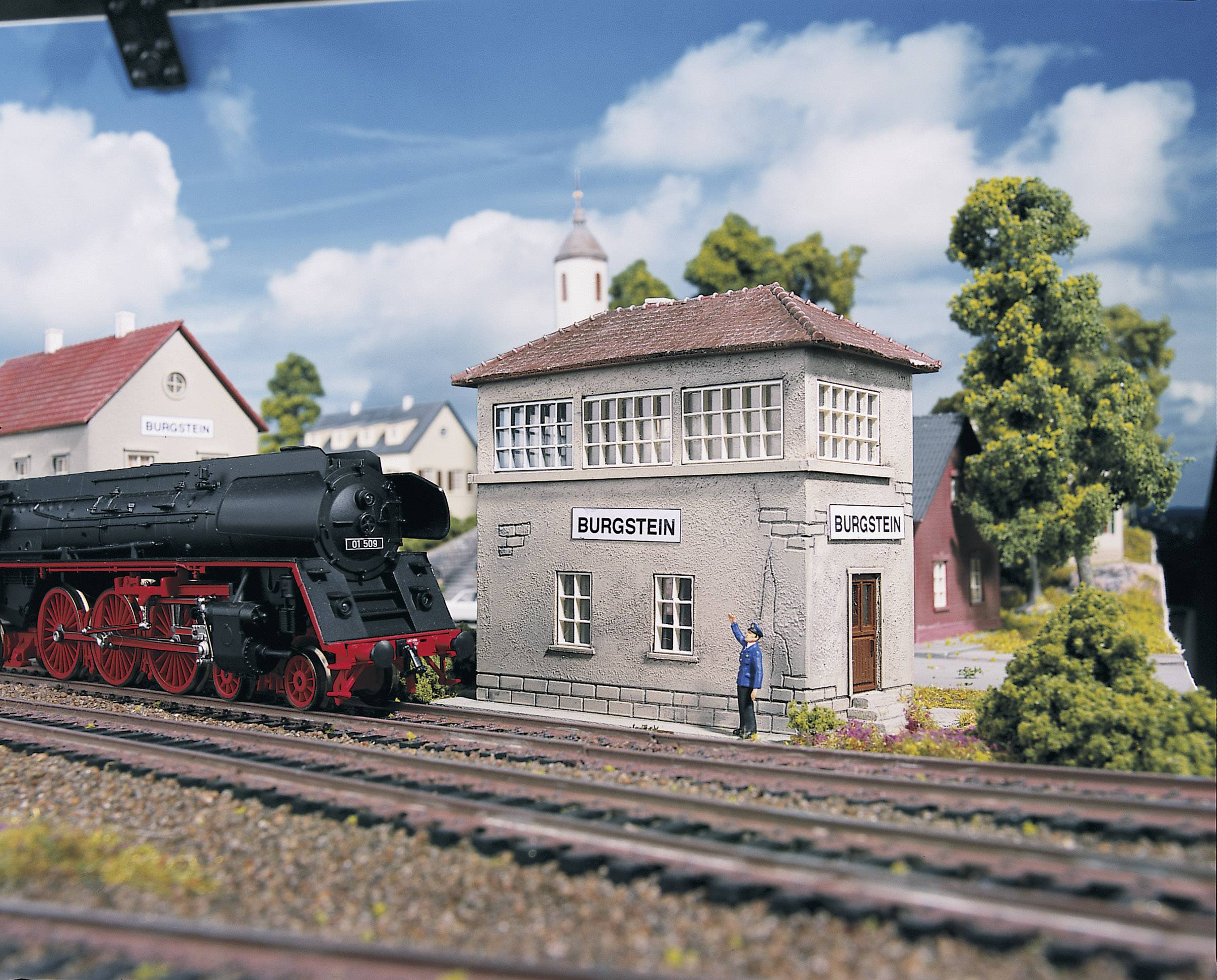 Un paysage miniature avec une locomotive à vapeur à côté d'un petit poste de signalisation portant l'inscription 'Burgstein', entouré d'arbres et de bâtiments.
