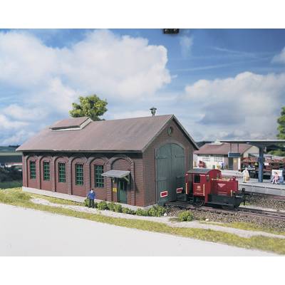Un modèle d'un ancien dépôt de locomotives avec une petite locomotive rouge devant. Des personnes se tiennent à proximité sur un quai.
