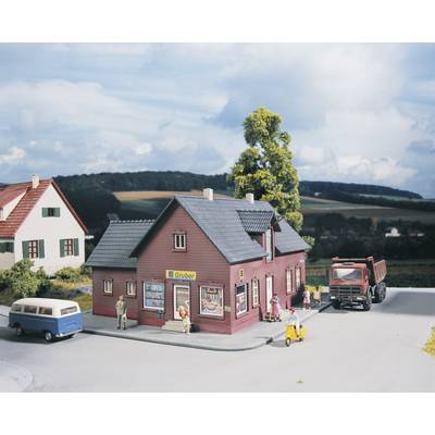 Un petit bâtiment de magasin de village rouge portant l'enseigne 'E. Gruber', entouré de personnes, d'un van bleu, d'un scooter et d'un camion.