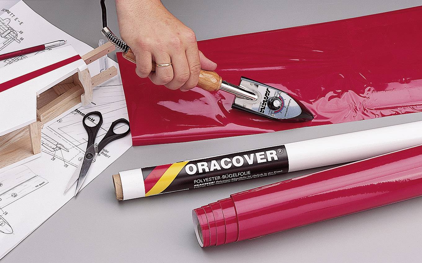 Une main utilise un petit fer à repasser pour coller un tissu en feuille de polyester rouge sur un modèle réduit d'avion. Des plans et des ciseaux sont posés en arrière-plan.