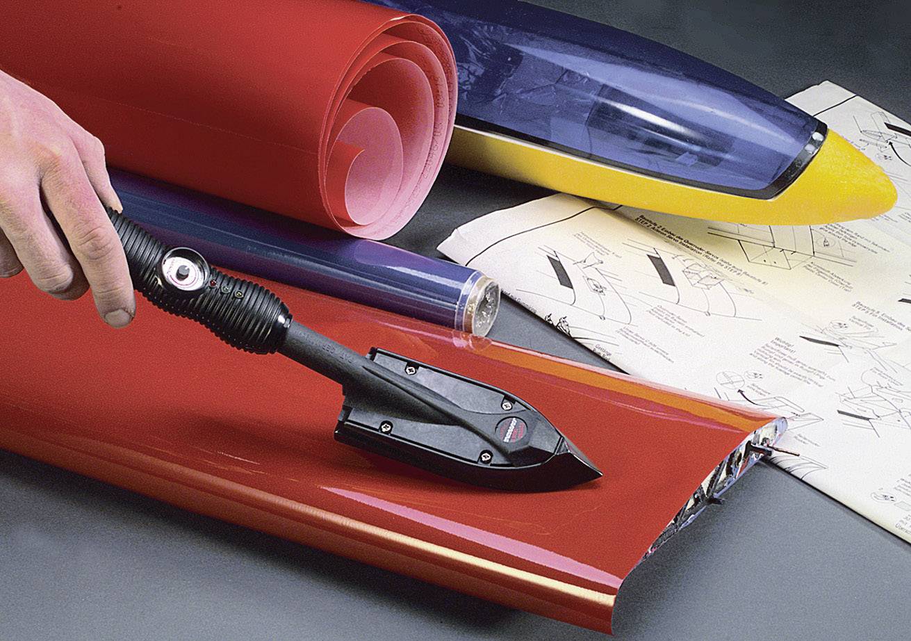 Une main tient un fer à repasser qui lisse une feuille de plastique rouge. En arrière-plan, des plans de construction d'un modèle d'avion et des rouleaux de feuilles de couleur sont visibles.