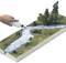 Une main verse du liquide d'une bouteille sur une maquette de paysage avec une rivière et des arbres pour représenter de l'eau.