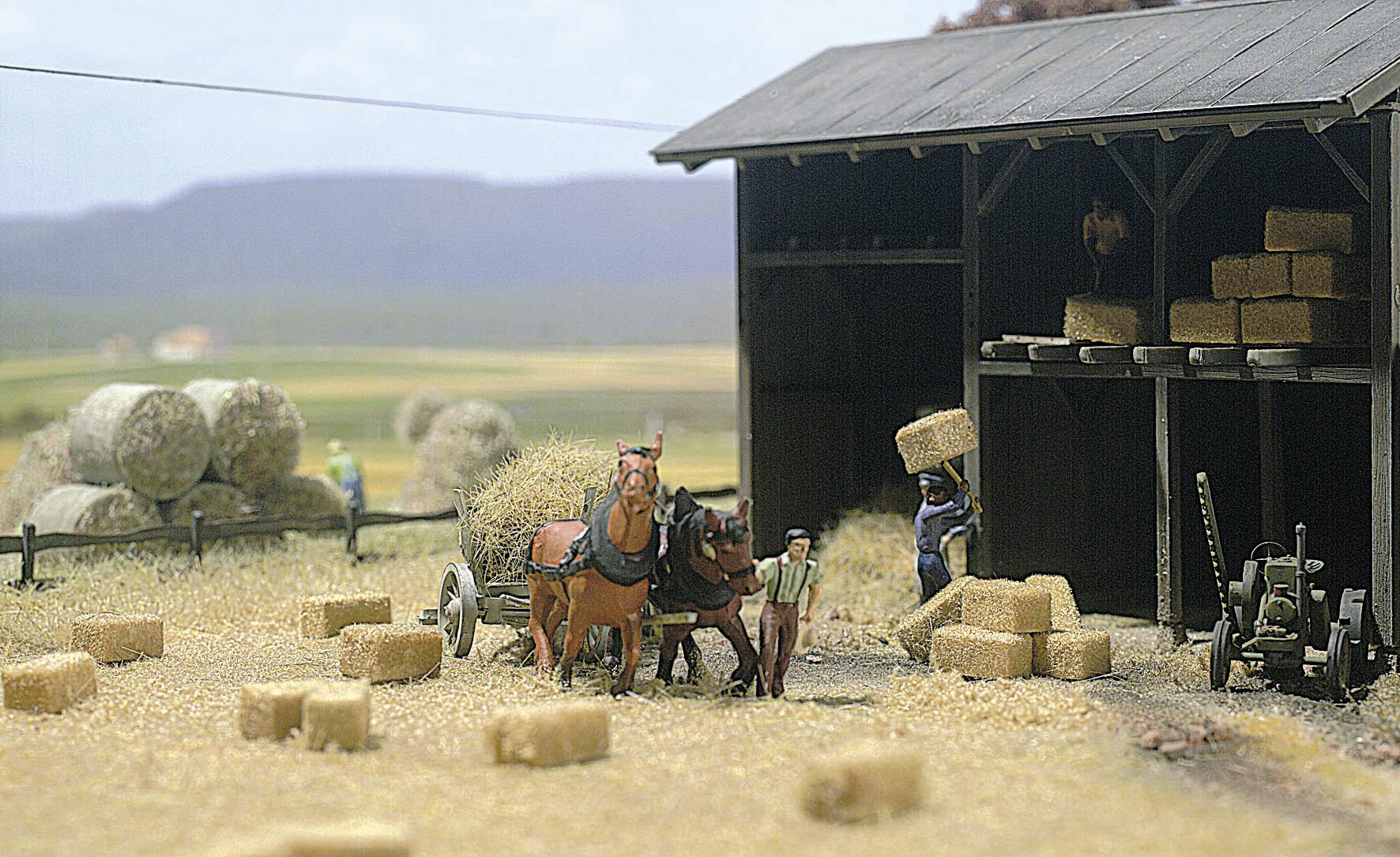 Des chevaux tirent une charrette de foin devant une grange. Deux personnes empilent des balles de foin. Des champs et des montagnes sont visibles en arrière-plan. Scène rurale.
