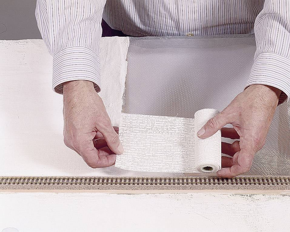 Une personne applique des bandes de plâtre sur un réseau de train miniature pour créer un paysage. La scène montre un travail pratique de modélisme.