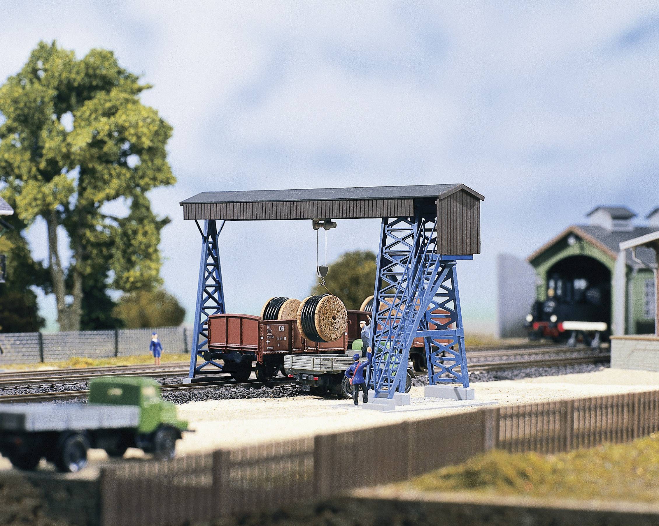 Un modèle de paysage ferroviaire représente un train de marchandises chargé de bobines de câbles, une grue et une locomotive en arrière-plan, le tout entouré d'arbres.