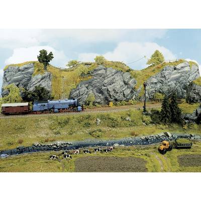 Un train miniature traverse un paysage rural avec des collines, des rochers et des prairies. Au premier plan, des vaches paissent.