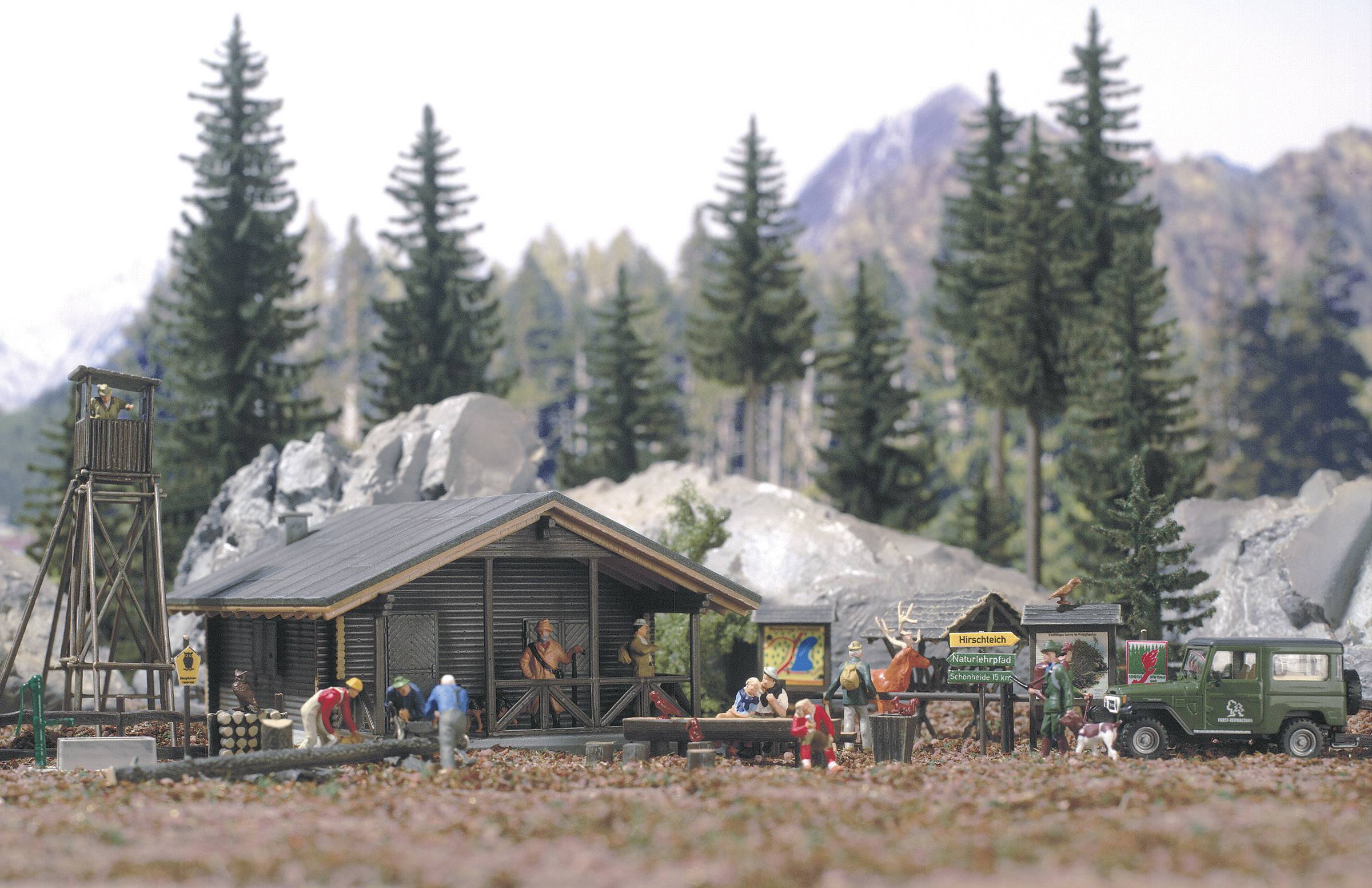 Petit village miniature dans un paysage montagneux : des personnes travaillent sur un chalet en bois, entourées d'arbres imposants et de rochers, avec des montagnes en arrière-plan.
