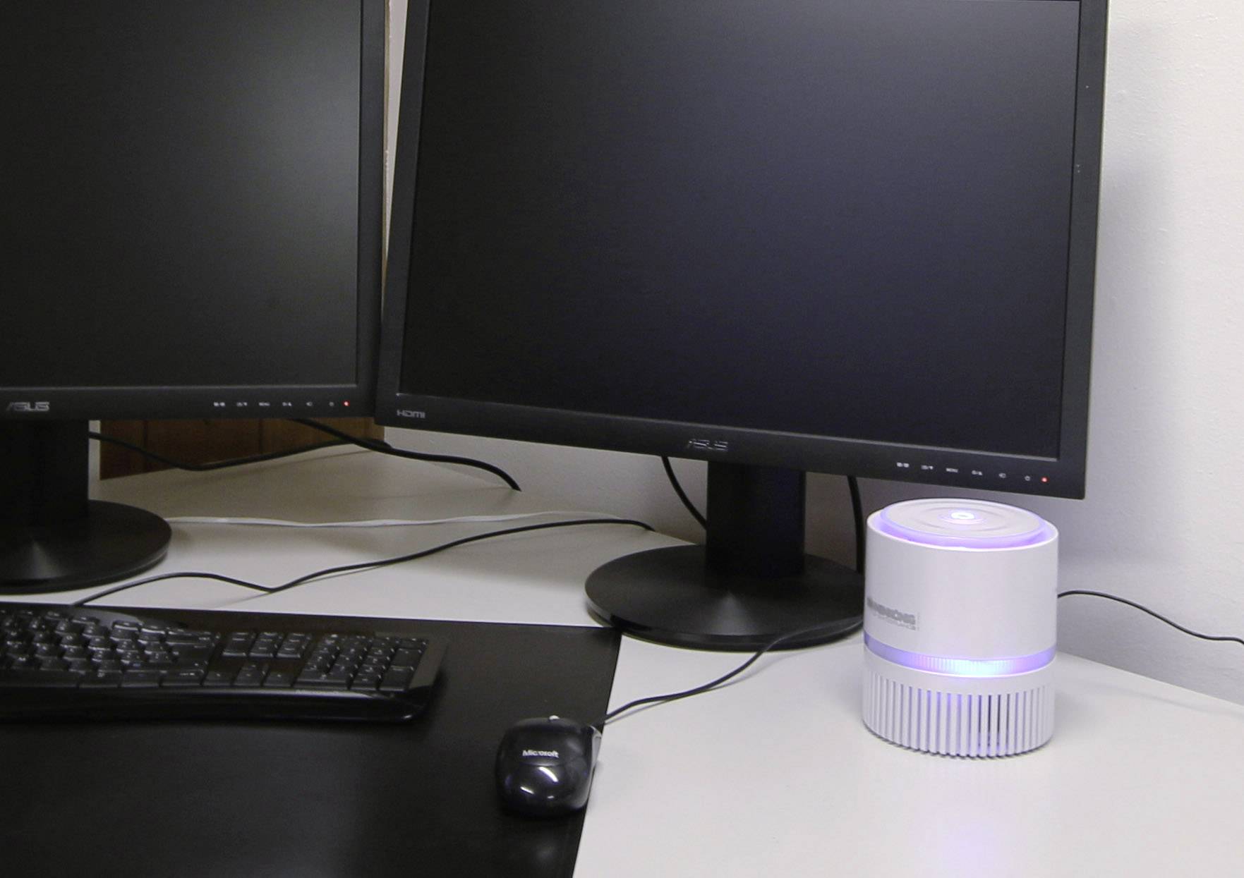 Bureau avec deux écrans, clavier et souris externes ; à côté, un purificateur d'air rond avec un indicateur lumineux.