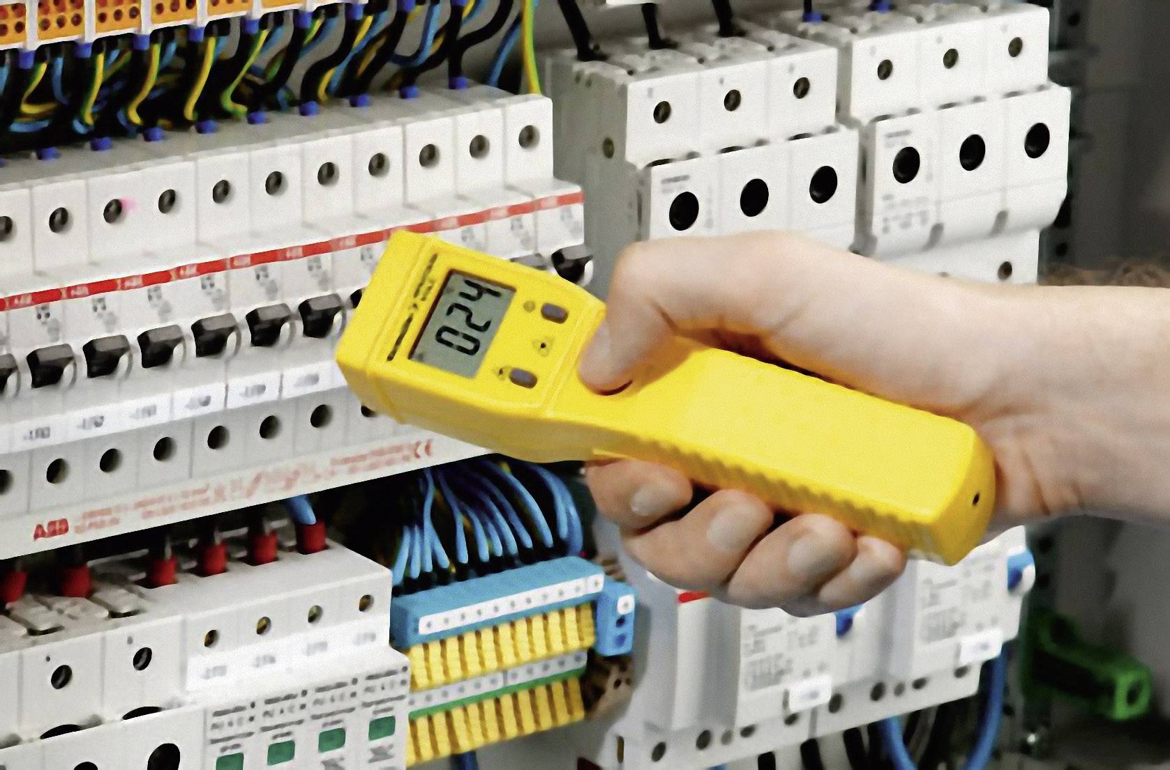 Une main tient un appareil de mesure numérique jaune devant un tableau électrique avec plusieurs interrupteurs et câbles.