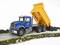 Un camion-jouet bleu avec une benne basculante jaune déverse des pierres sur une route. Le camion est détaillé et se trouve sur un fond vert.