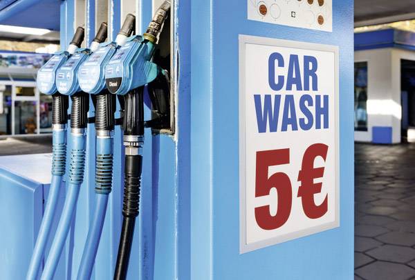 Une pompe à essence avec plusieurs pistolets bleus. Un panneau sur la colonne propose un lavage de voiture pour 5 EUR.