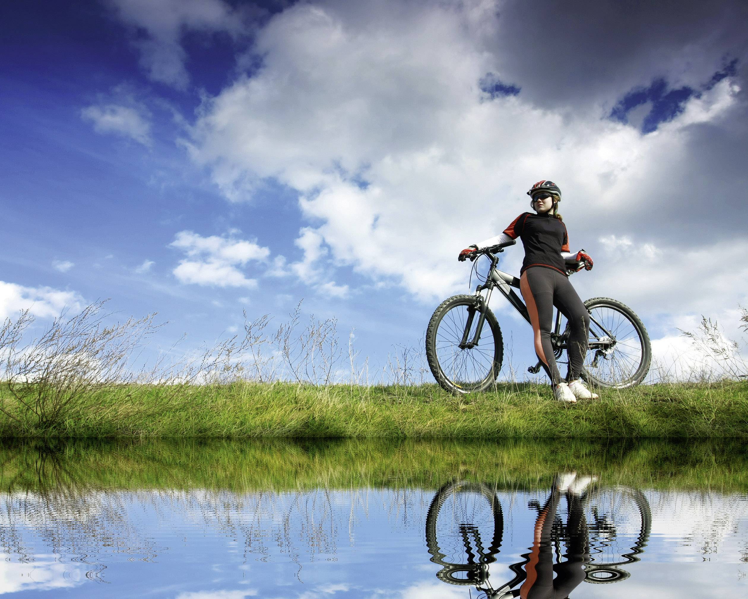 Une personne se tient avec son vélo au bord d'un plan d'eau sous un ciel bleu parsemé de nuages. L'image se reflète dans l'eau.
