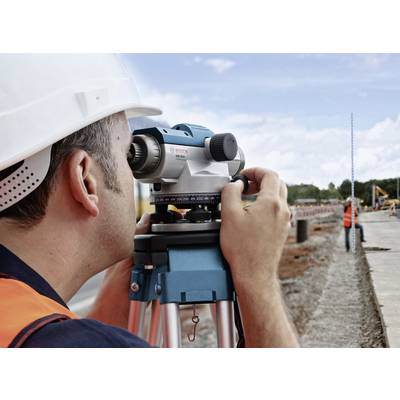 Une personne portant un casque de protection recherche un point sur un chantier à l'aide d'un niveau de chantier. Des travaux routiers sont visibles en arrière-plan.