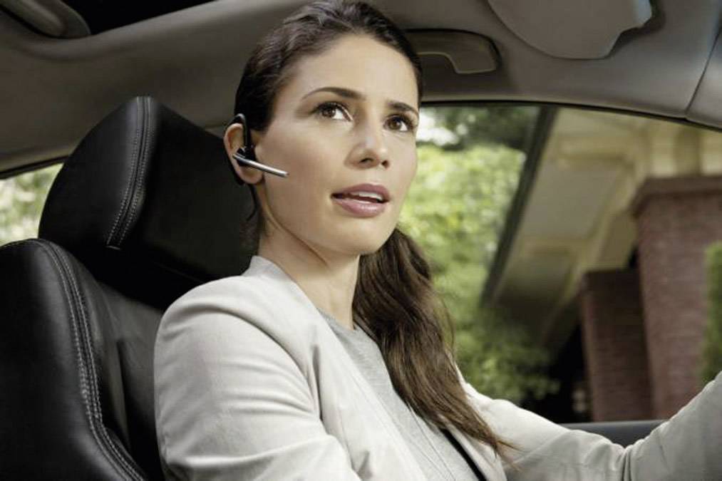 Une femme est assise dans une voiture et parle dans un casque. Elle porte un blazer clair. Une maison est visible en arrière-plan.