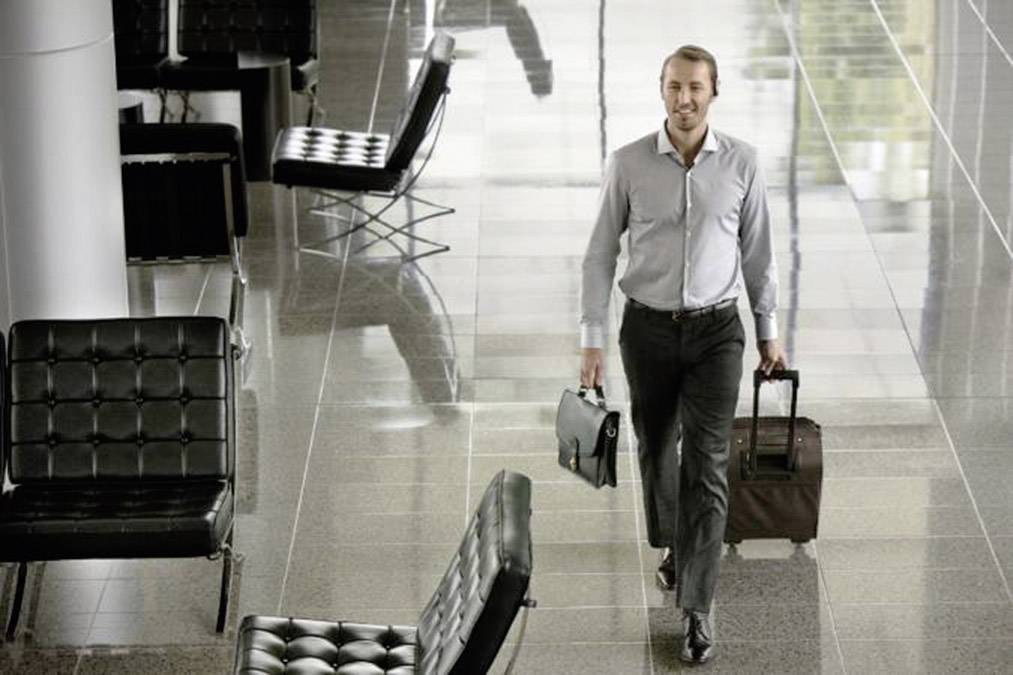 Un homme en costume d'affaires traverse un aéroport moderne, portant un attaché-case d'une main et une valise à roulettes de l'autre.