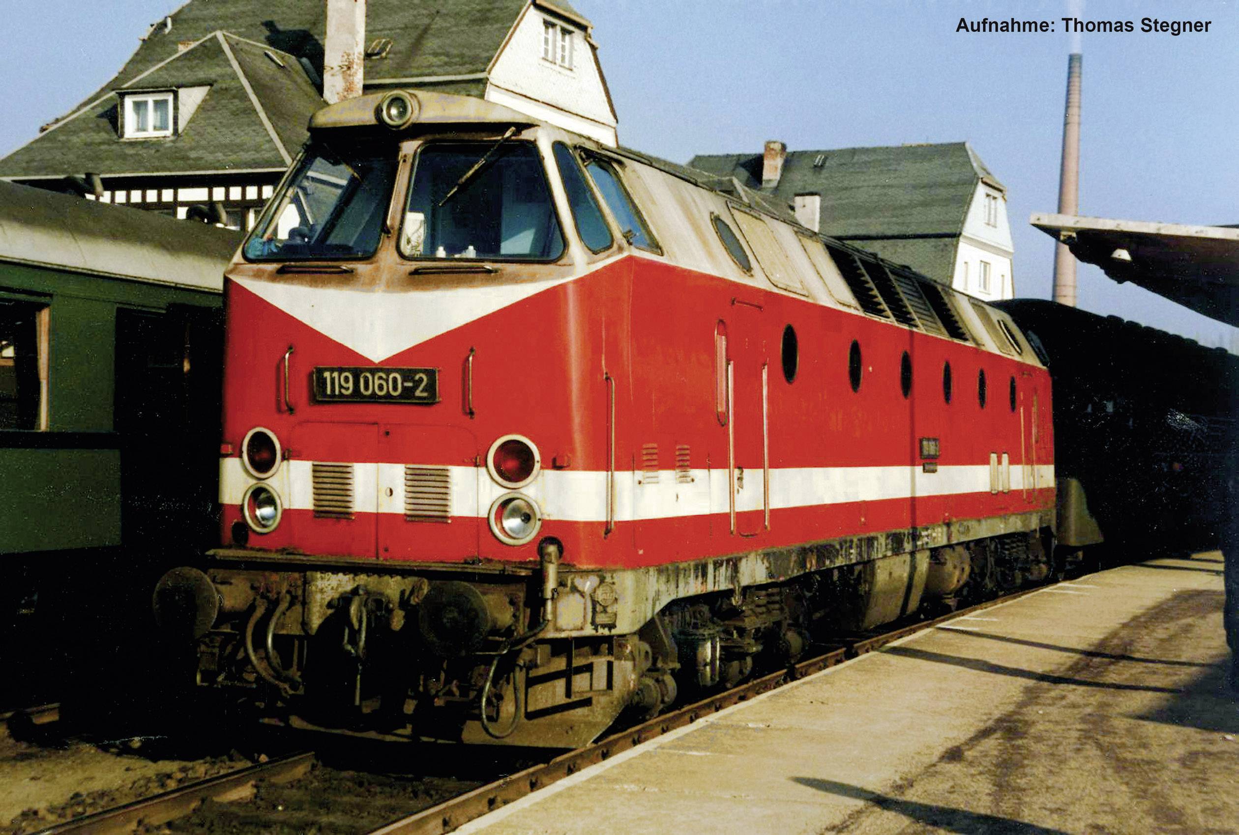 Une locomotive rouge de la série 119 est stationnée sur une voie ferrée à côté d'un wagon vert. Des bâtiments sont visibles en arrière-plan.