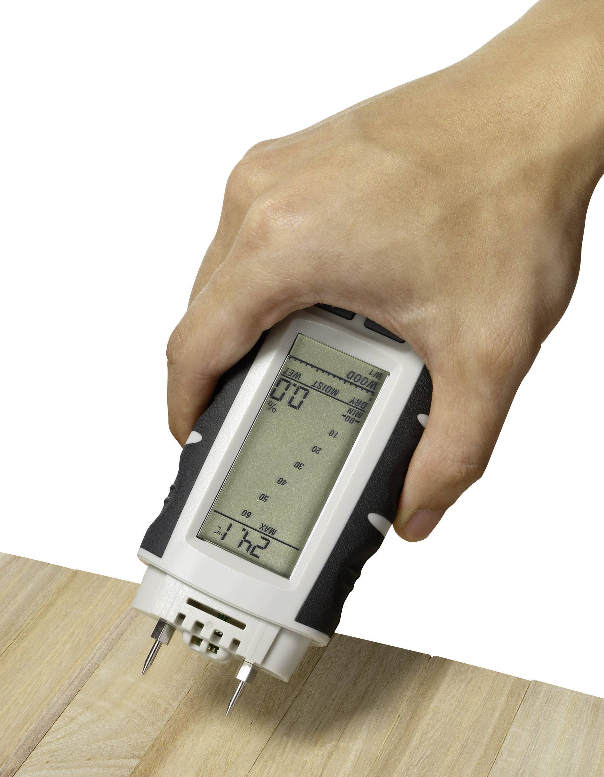 Une main tient un hygromètre numérique contre une planche de bois. L'appareil affiche des valeurs de mesure sur un écran.