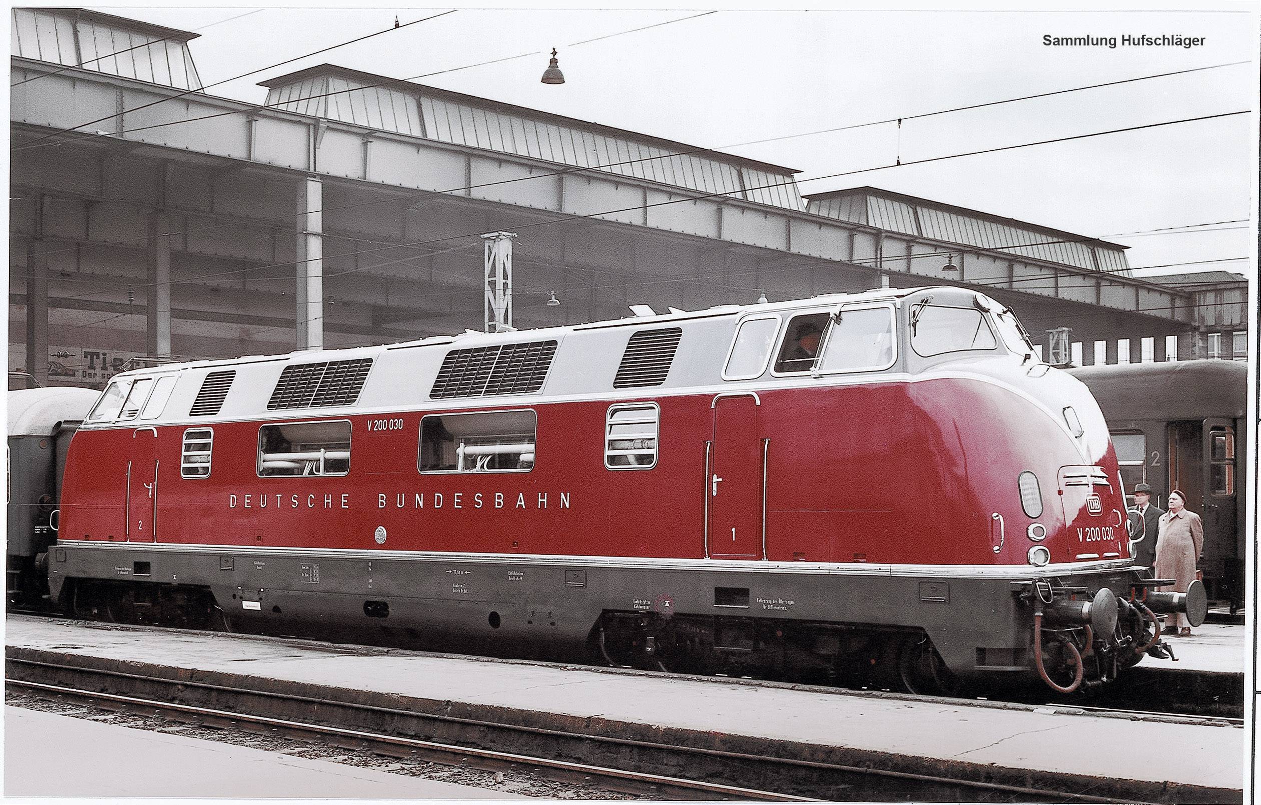 Un train de locomotives diesel rouges portant l'inscription 'Deutsche Bundesbahn' est stationné sur une voie de gare. Des bâtiments et des personnes sont visibles en arrière-plan.
