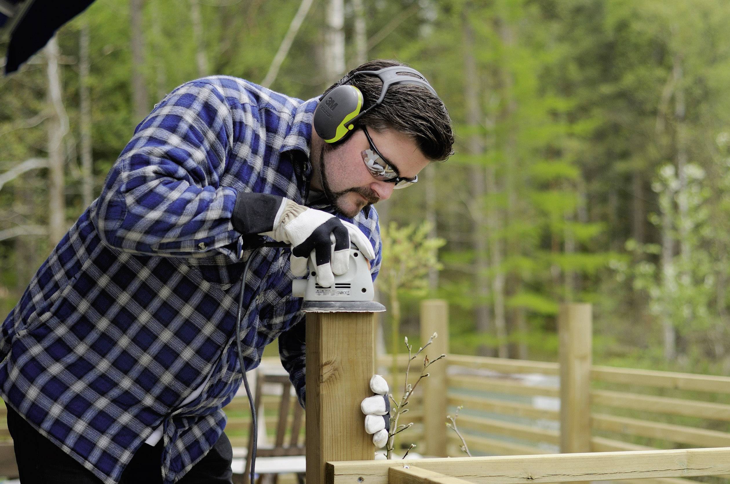 Un homme portant un équipement de protection utilise une meuleuse électrique pour travailler un poteau extérieur en bois dans un environnement boisé.