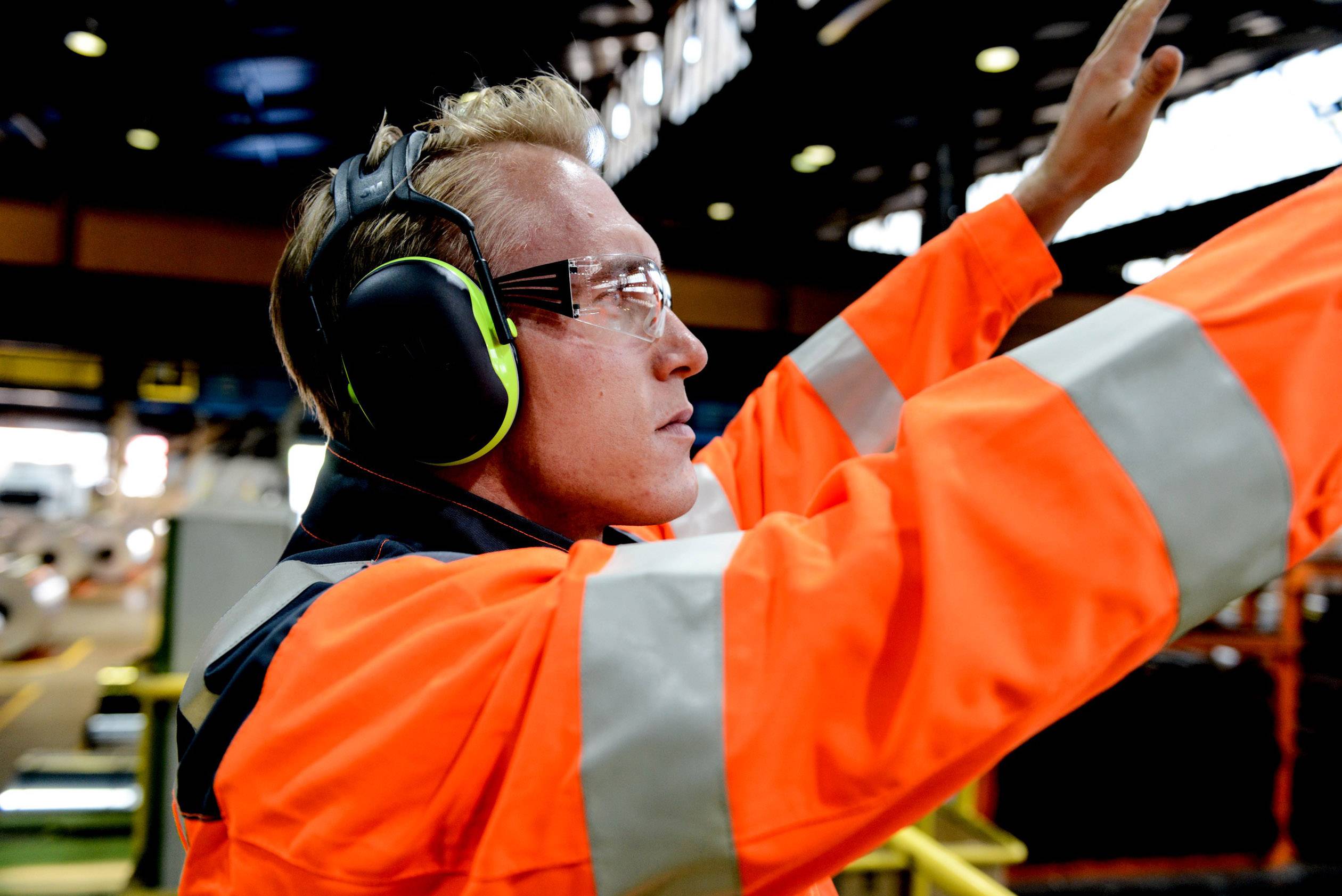 Un travailleur portant un équipement de protection orange et des protections auditives lève un bras, probablement pour donner un signal dans un environnement industriel.