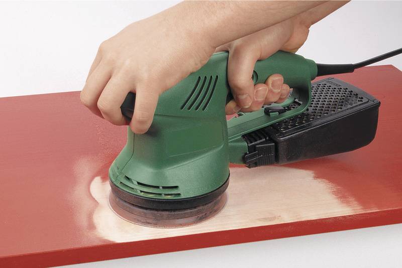 Une main tient une ponceuse excentrique verte qui ponce la peinture rouge d'une planche en bois.