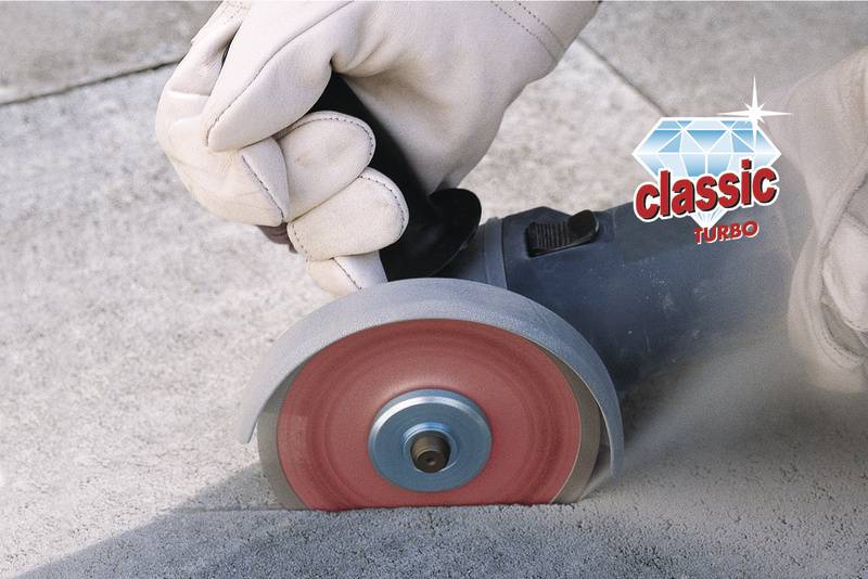 Un meuleuse découpe du béton avec un disque rouge. Une main gantée tient l'outil. La marque 'classic TURBO' est visible.