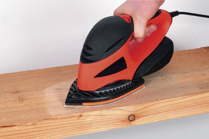 Une main tient une ponceuse orbitale rouge qui est utilisée sur une planche en bois.