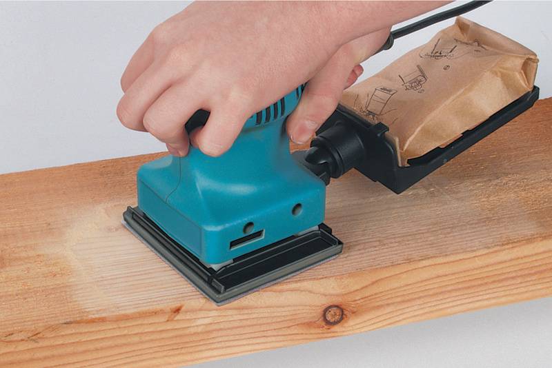 Une main tient une ponceuse électrique qui est déplacée sur une planche de bois pour en lisser la surface.