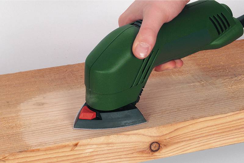 Une main tient un outil de ponçage vert qui glisse sur une planche en bois pour en lisser la surface.