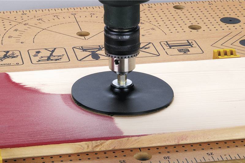 Perceuse à main avec un disque de meulage ponce une planche de bois peinte en rouge sur l'établi. Diverses échelles de mesure sont visibles.