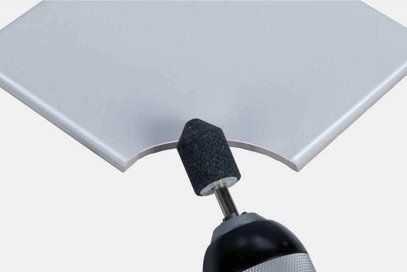 Un foret équipé d'un accessoire de meulage forme un bord incurvé sur un carreau blanc.