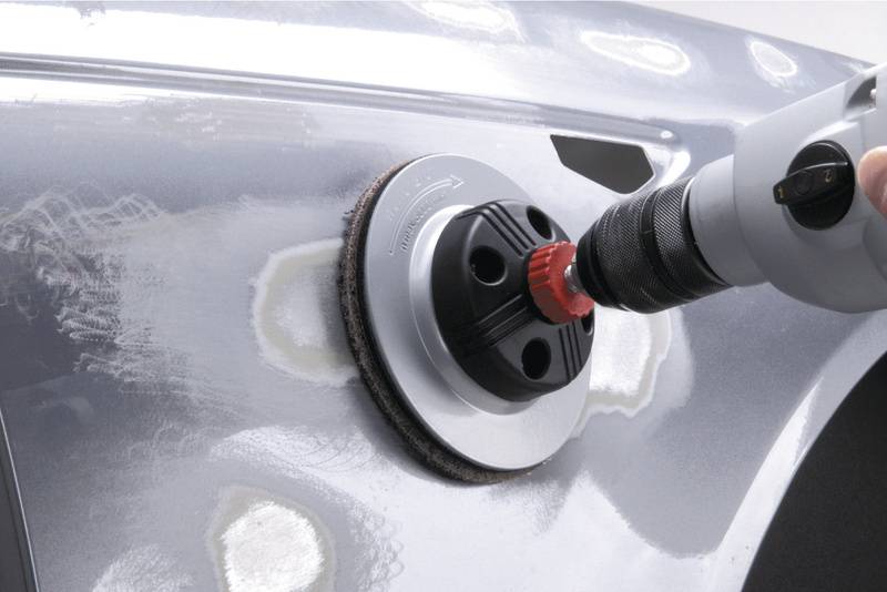 Une main tient une polisseuse qui travaille sur une surface de carrosserie rayée d'une voiture, afin de lisser la surface.