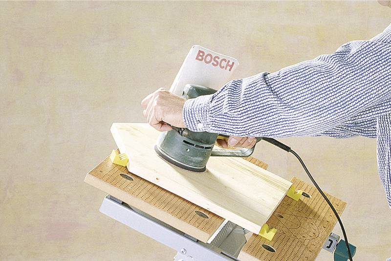 Une personne ponce une planche en bois avec une ponceuse Bosch sur une table de travail.
