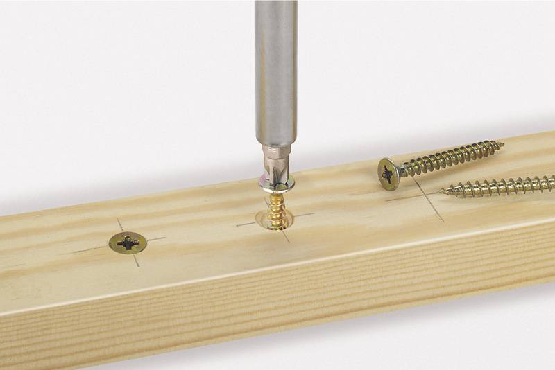 Un tournevis enfonce une vis dorée dans une planche de bois. Deux autres vis sont visibles sur la planche.