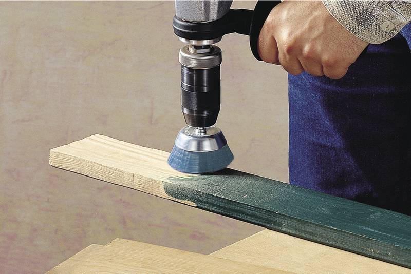 Une main tient une perceuse équipée d'une brosse métallique pour poncer la peinture verte d'une planche en bois.