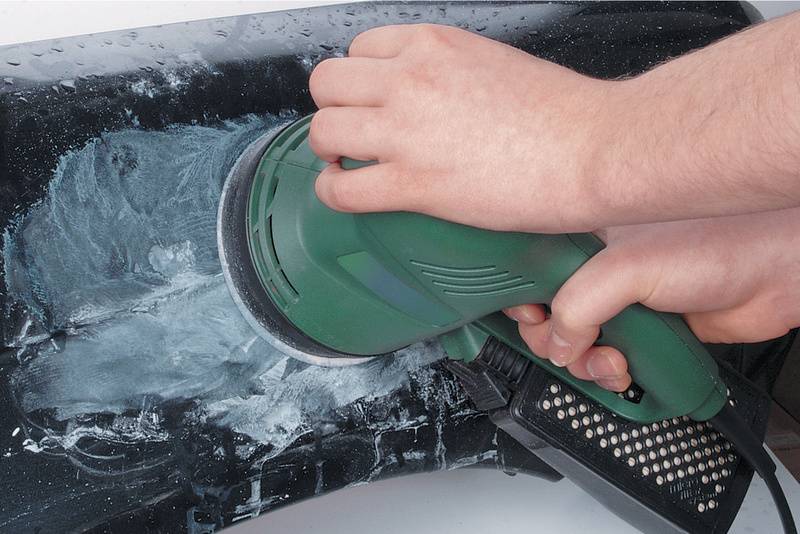 Deux mains tiennent une meuleuse électrique et meulent un objet noir sur lequel on peut voir de la poussière blanche de ponçage.