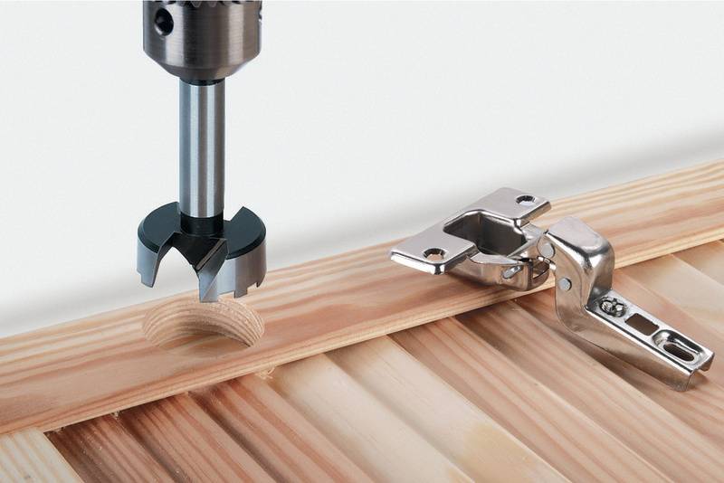 Une perceuse travaille une planche en bois pour la préparation du montage d'une charnière, qui est posée à côté.