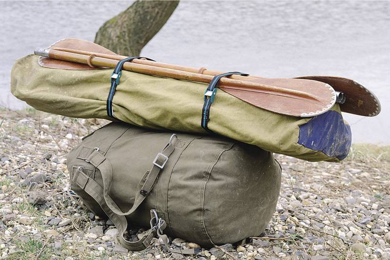 Un sac marin vert olive est posé sur un sol pierreux au bord de la rivière. Un tente pliée et deux pagaies en bois sont attachées sur le dessus.