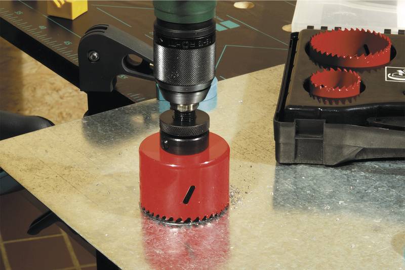 Une scie cloche rouge est montée sur une pièce de métal, prête à percer. À côté se trouve une mallette contenant d'autres lames de scie.