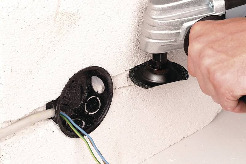 Une main tient un outil qui découpe une ouverture circulaire dans un mur blanc, à côté d'une prise électrique murale déjà existante.