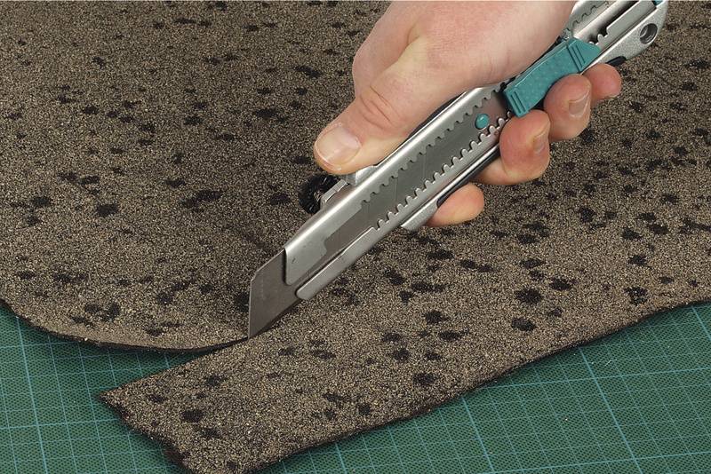 Une main utilise un couteau à tapis pour couper précisément un tapis brun avec des taches noires sur un tapis de coupe vert.
