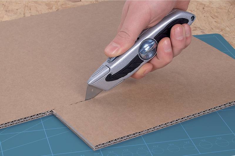 Une main coupe un carton avec un cutter argenté sur un tapis de coupe vert.