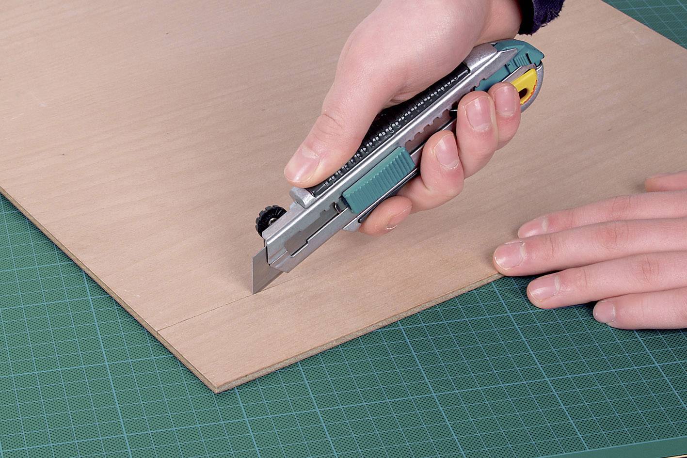 Une main tient un cutter et coupe un morceau de contreplaqué sur un tapis de coupe vert.