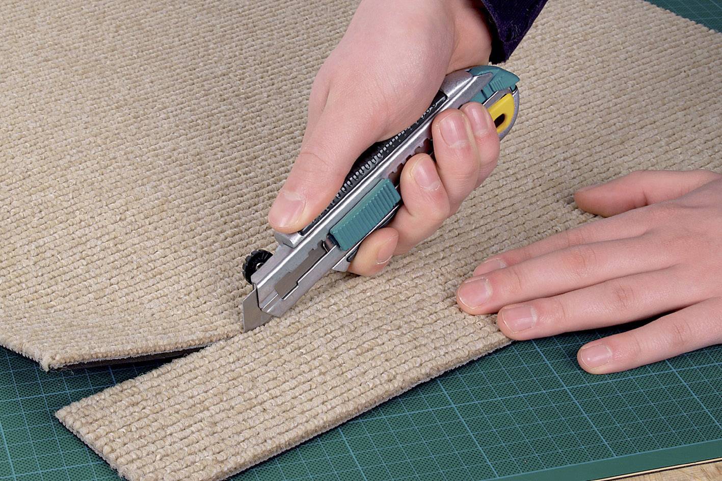 Une personne coupe un tapis beige avec un couteau de tapissier sur un tapis de coupe vert.